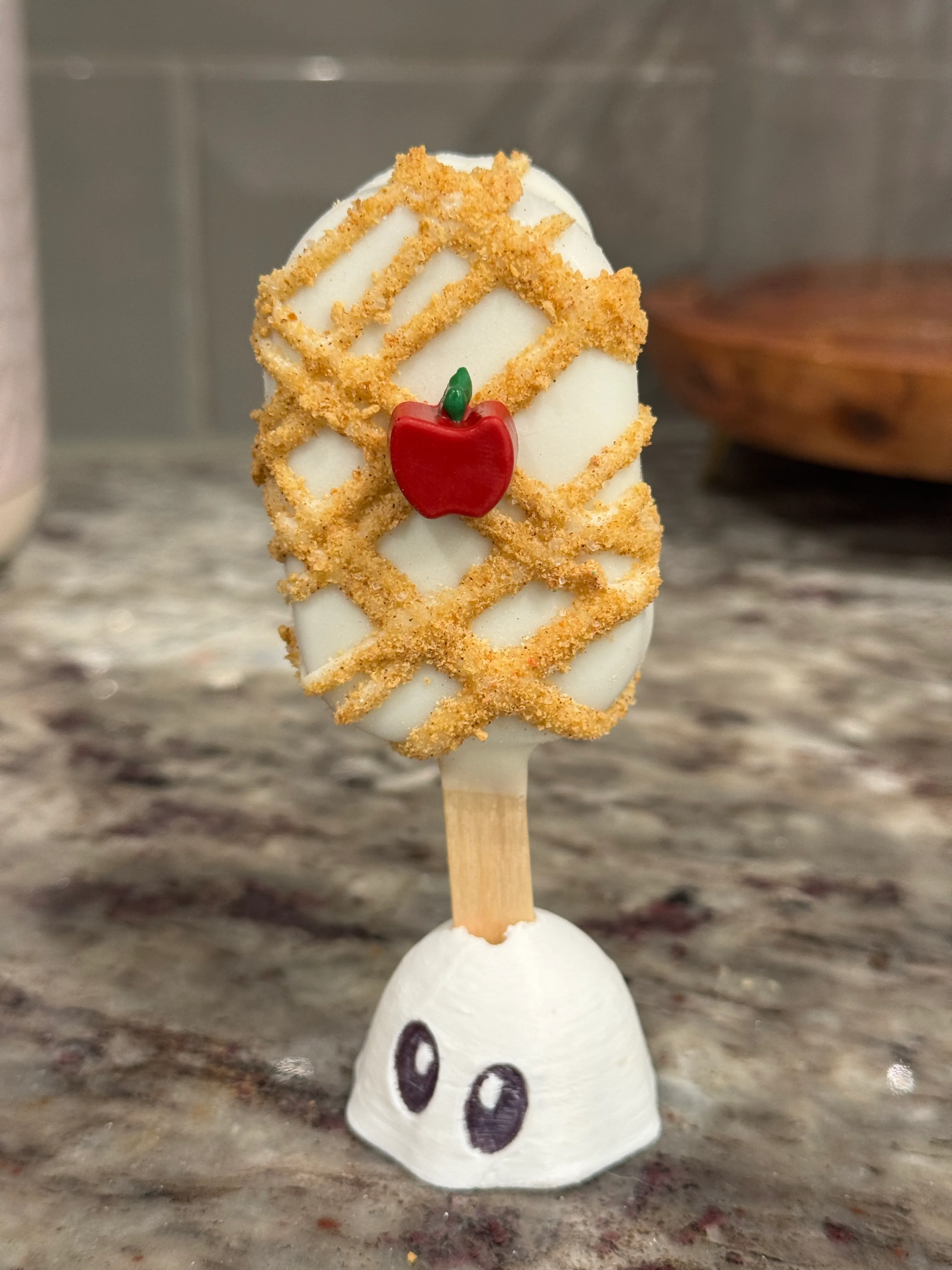 Apple pie cider cakesicle in a white shell with a lattice drizzle and cinnamon crumb, topped with a red fondant apple