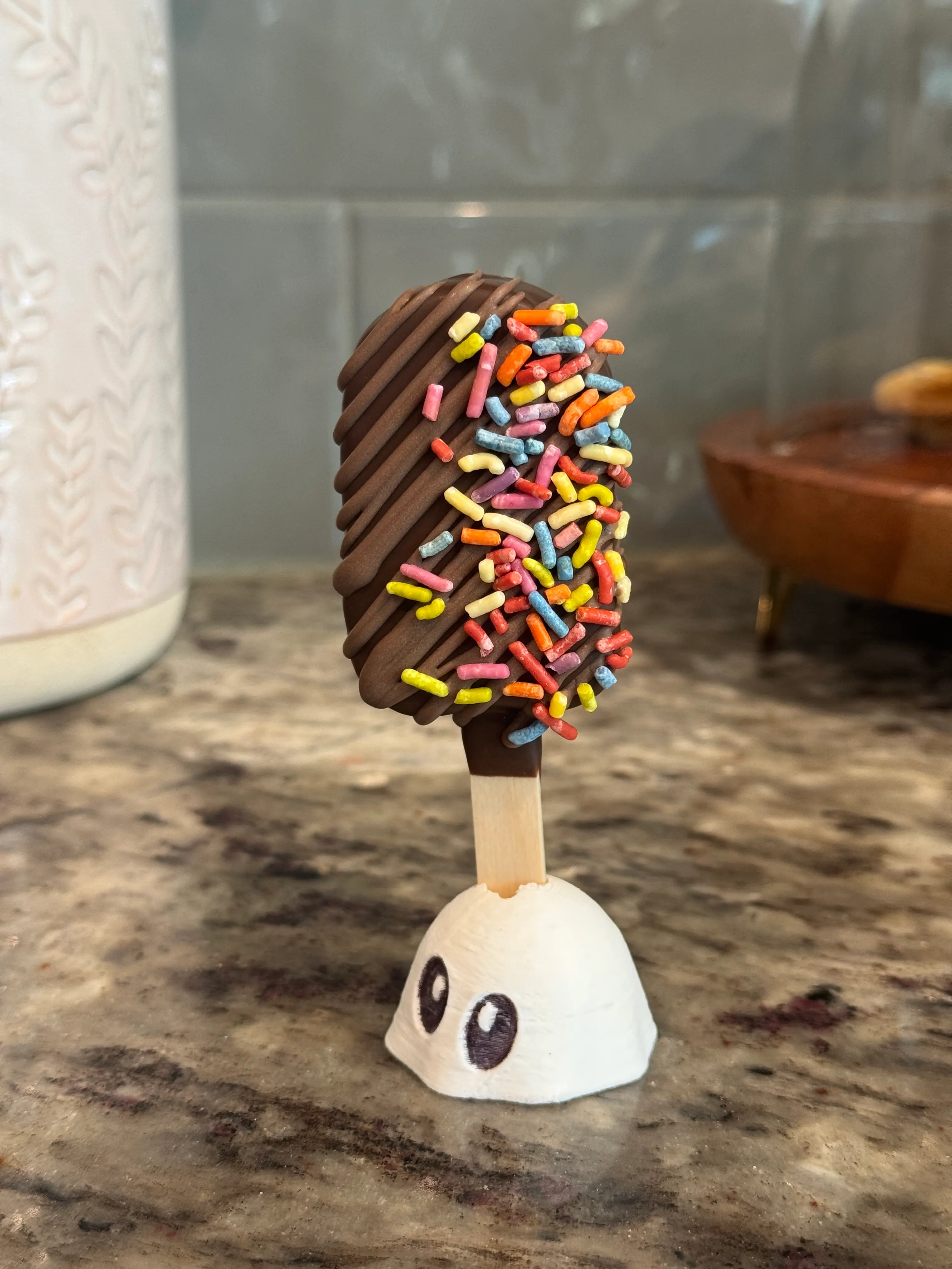Gluten-free chocolate funfetti cakesicle with a chocolate drizzle and rainbow sprinkles, standing in a ghost holder