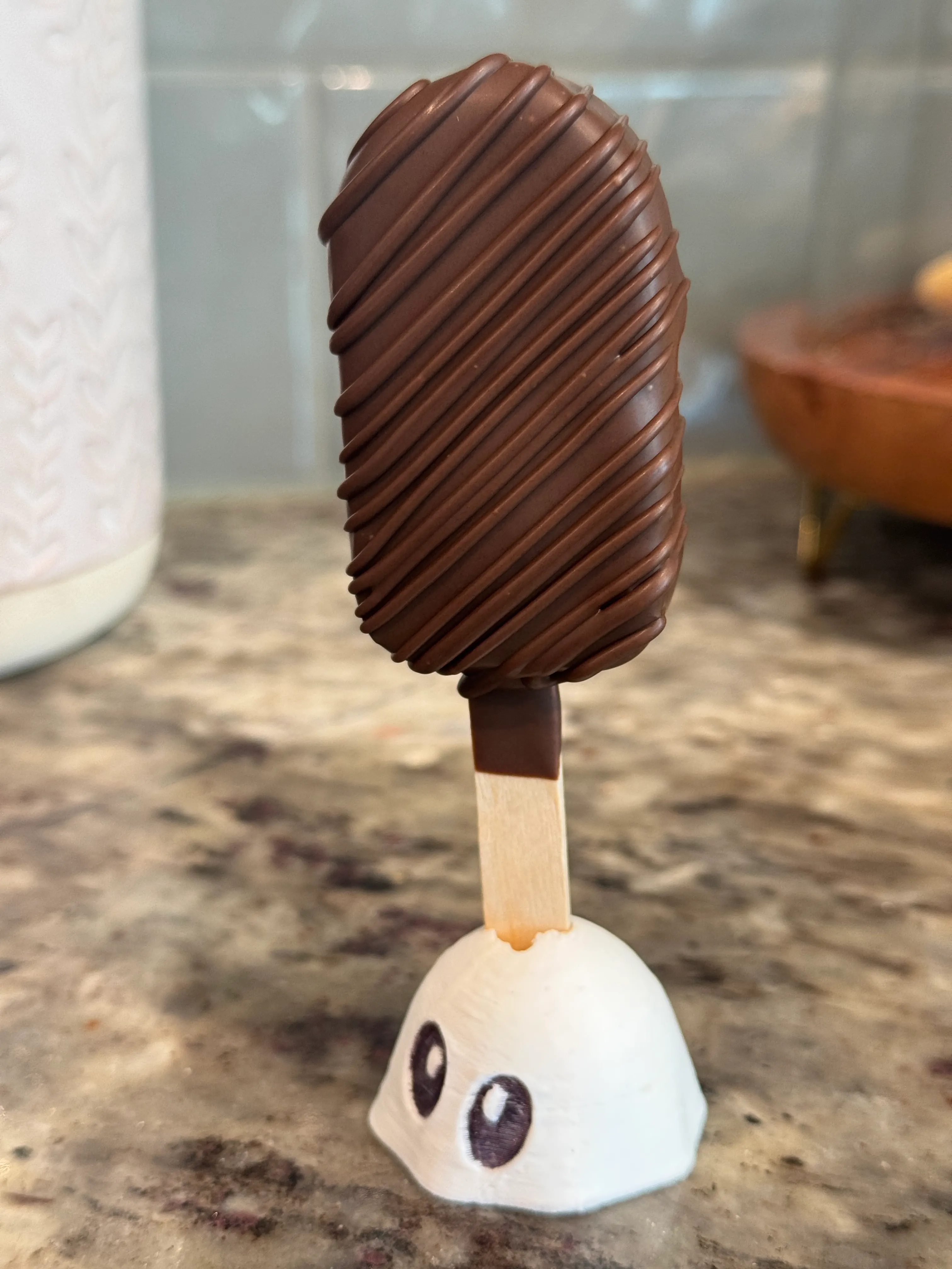 Gluten-free chocolate cakesicle in a plain chocolate shell with a ribbed drizzle, standing in a ghost holder