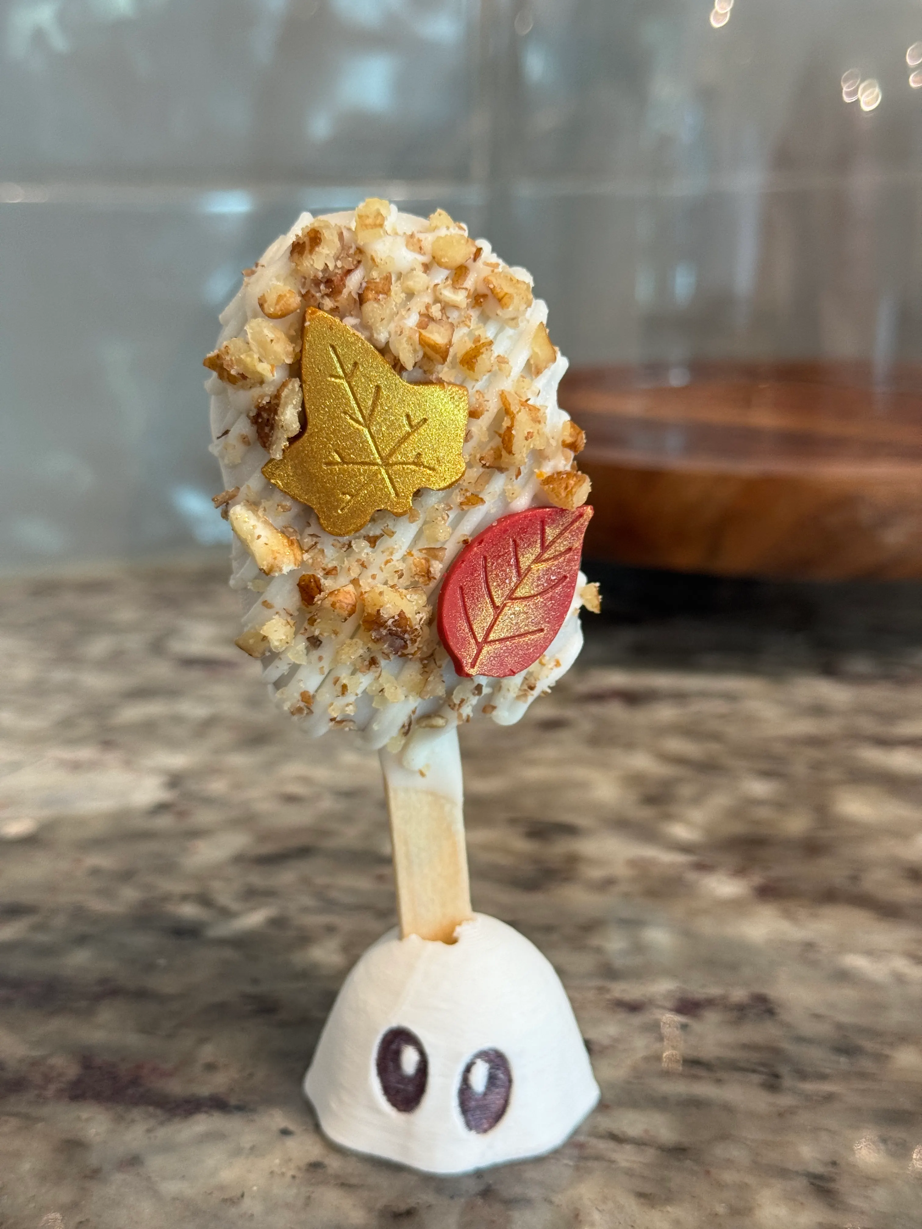 Maple butter pecan cakesicle in a white shell with white drizzle, chopped pecans and gold and red fondant leaves