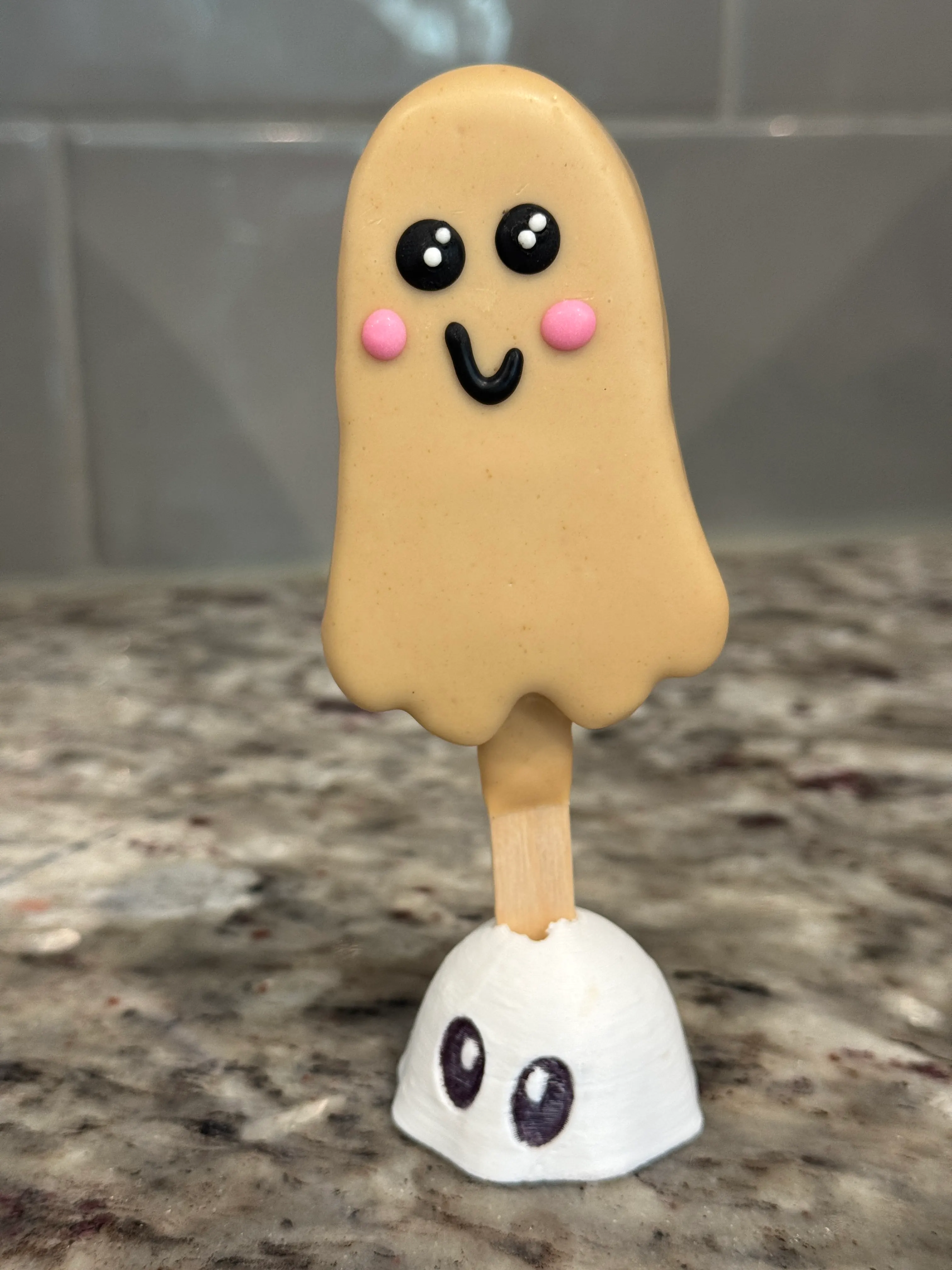 Boo-scoff cakesicle shaped like a ghost in a tan cookie butter shell, with black candy eyes, a smile and pink cheeks