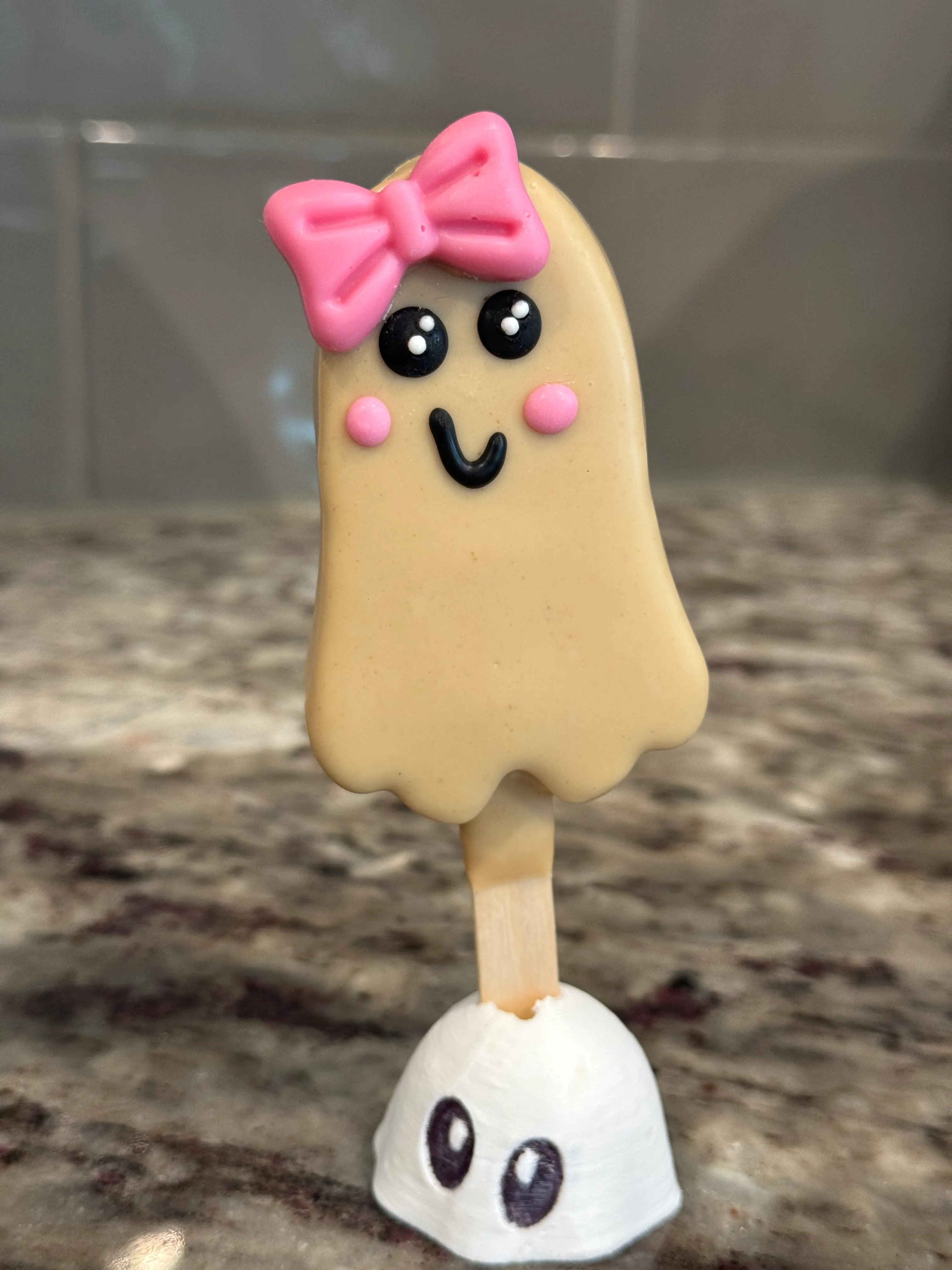 Boo-scoff ghost cakesicle in a tan cookie butter shell wearing a pink bow, with black candy eyes and pink cheeks