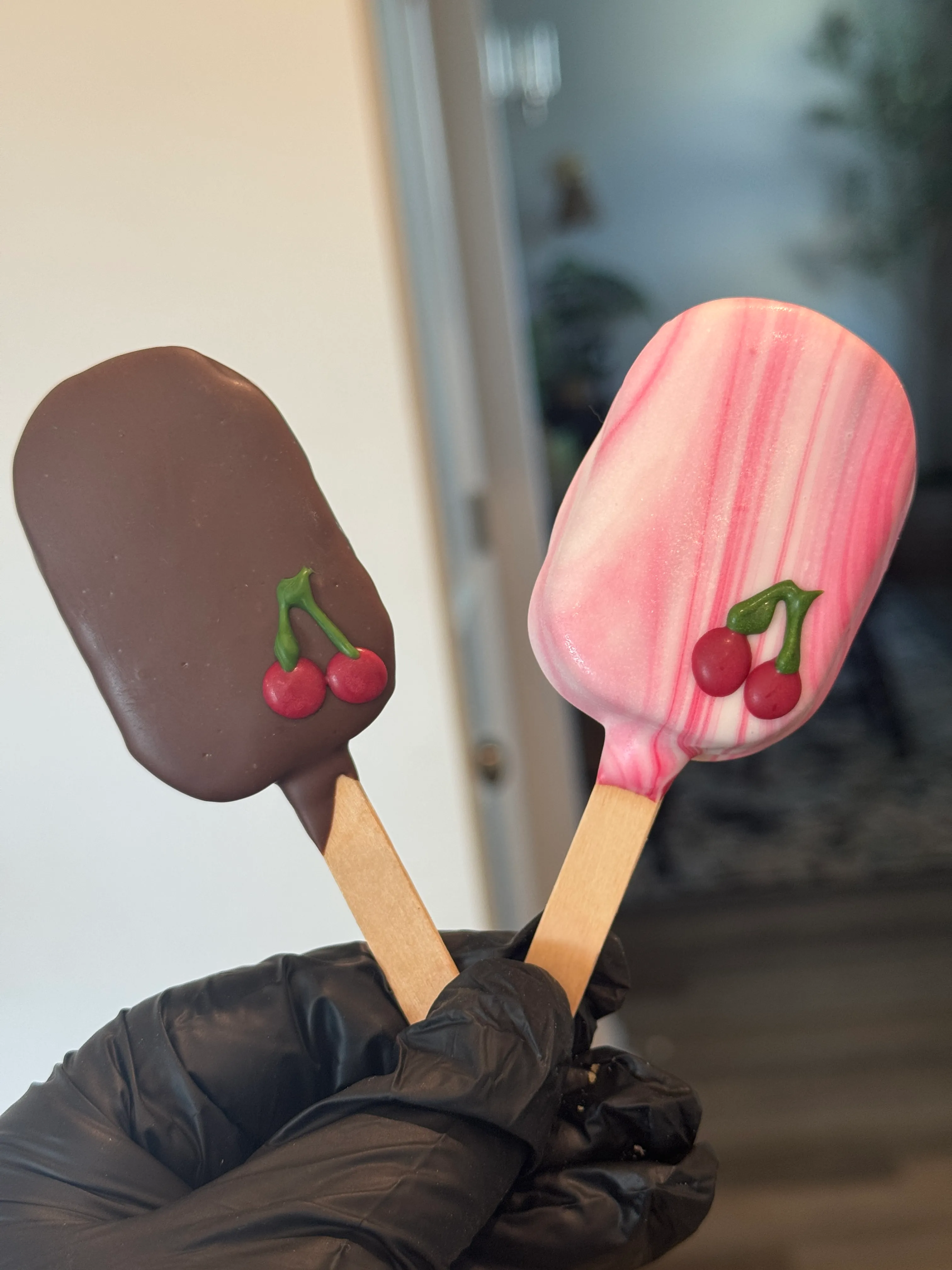 A pink marbled cherry pie cakesicle beside a dark chocolate cherry cakesicle, each piped with two cherries
