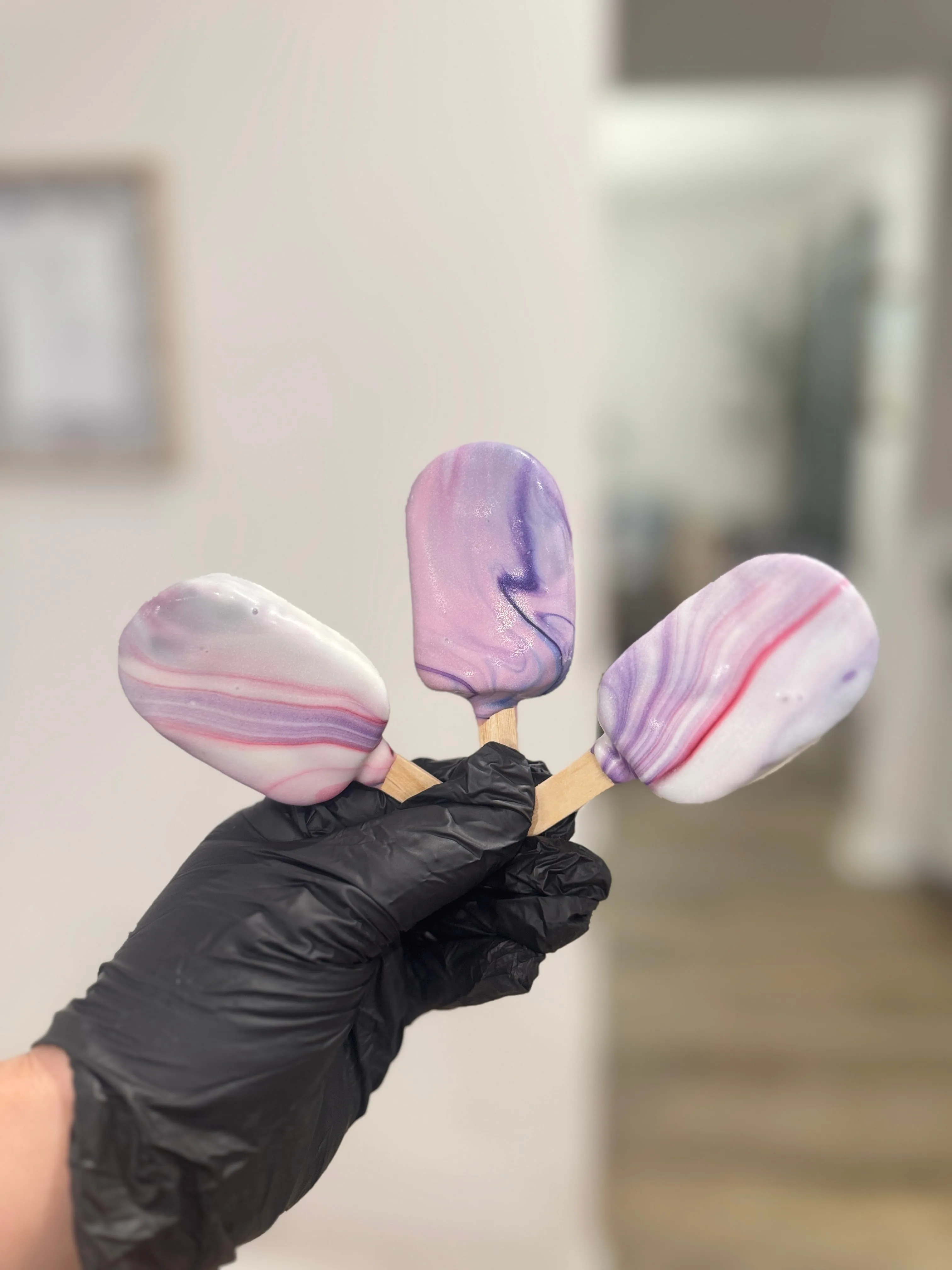 Three marbled cakesicles in pink and purple swirls held up in a gloved hand