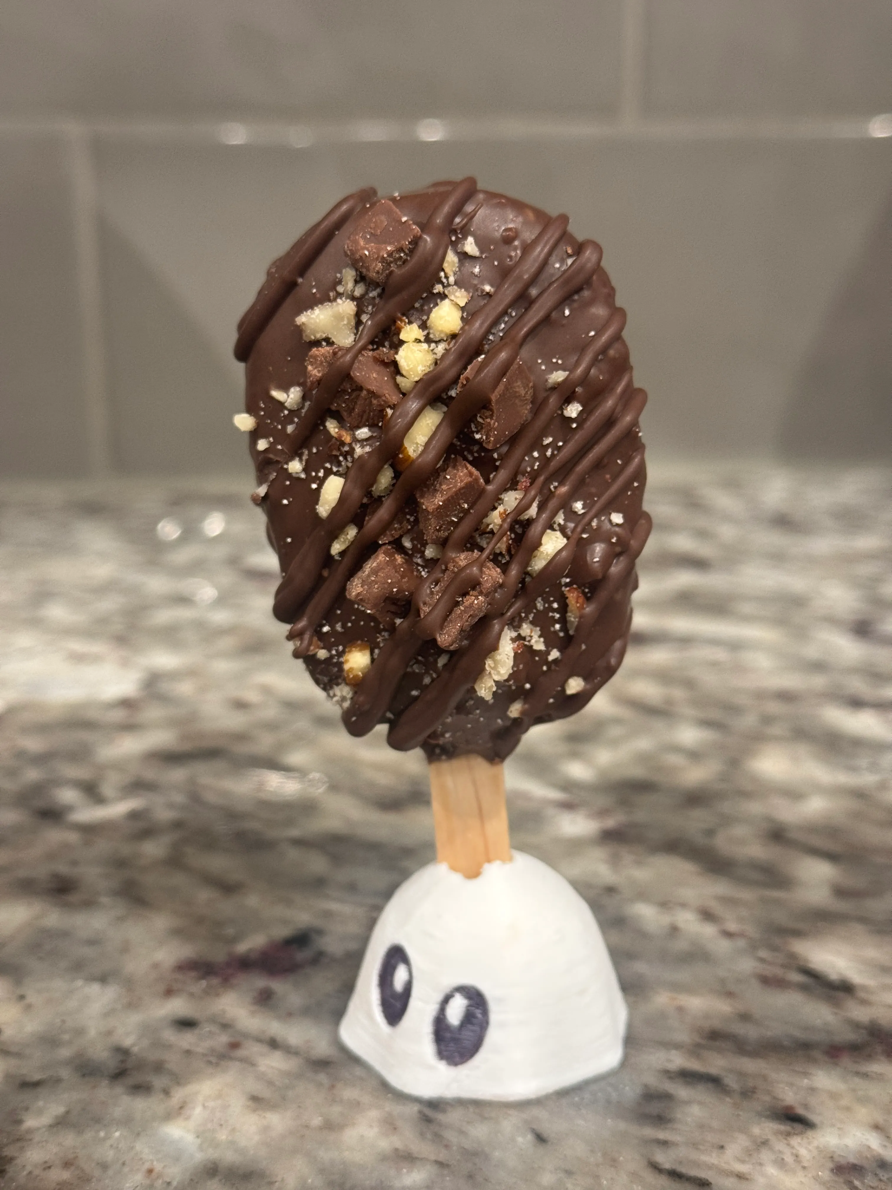 Hazelnut truffle cakesicle in a dark chocolate shell, topped with chopped hazelnuts and milk chocolate chunks under a chocolate drizzle