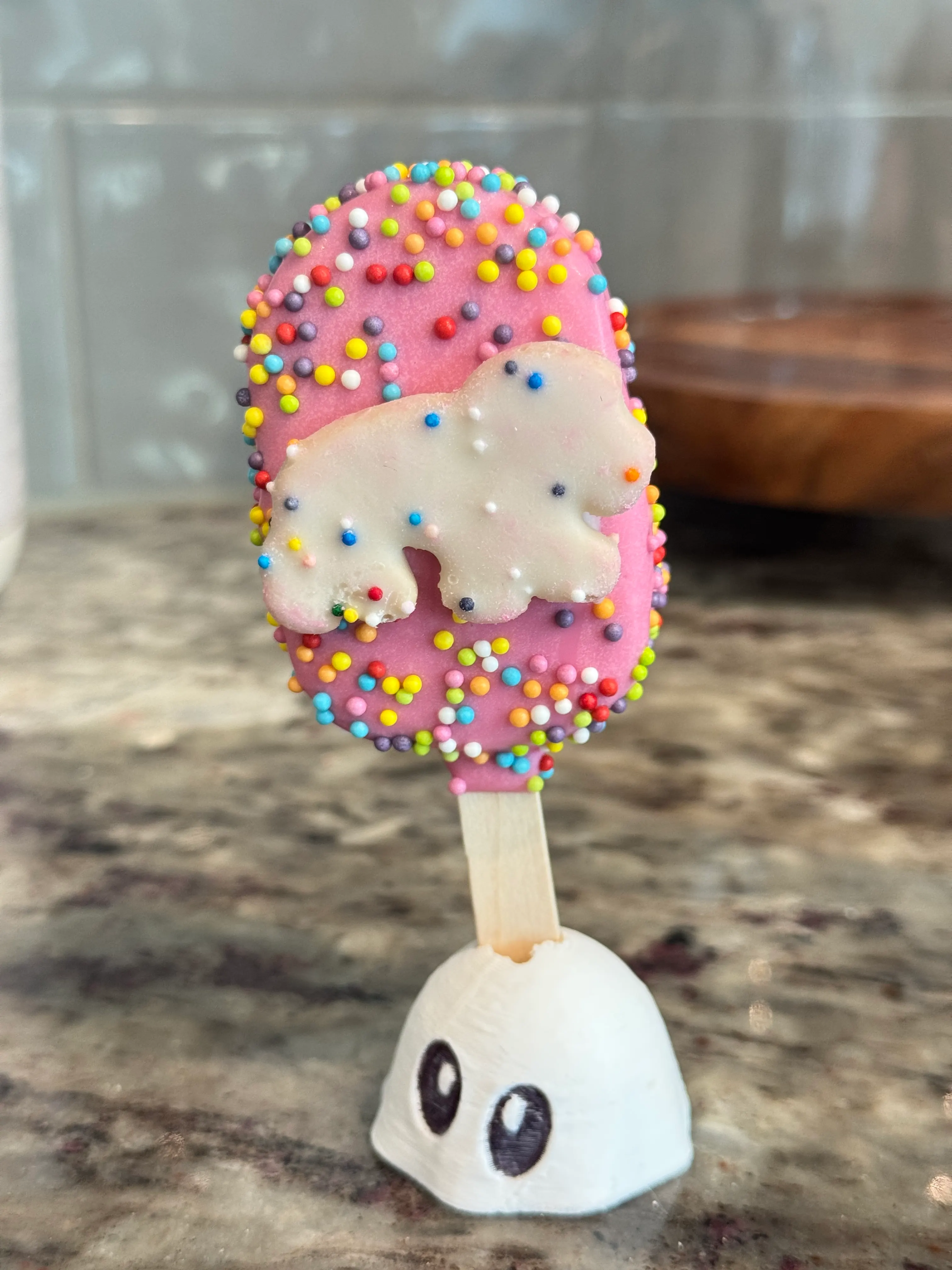 Circus cookie cakesicle in a pink shell covered in rainbow nonpareils, topped with a white circus animal cookie