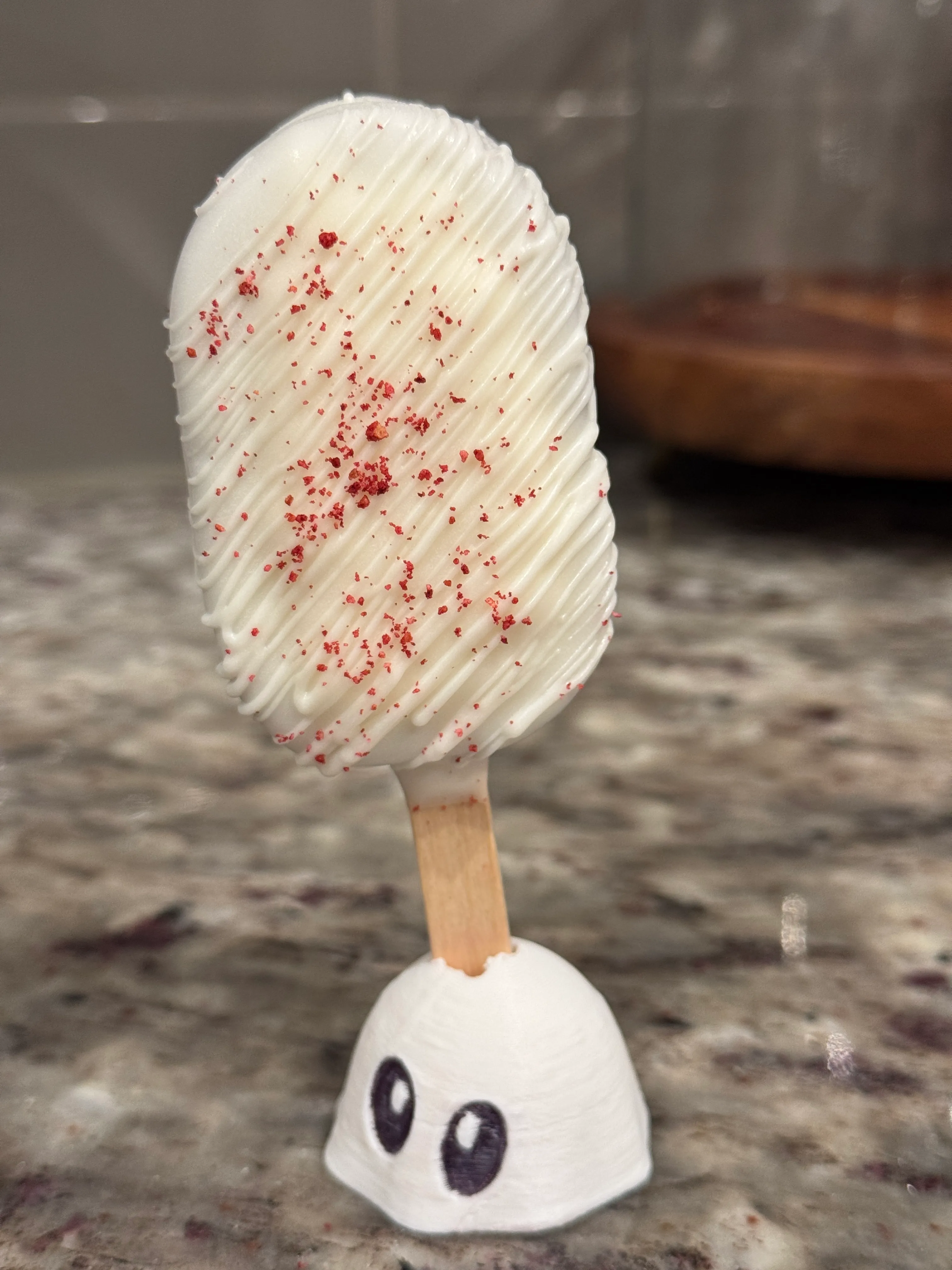 Red velvet cakesicle in a white shell with white drizzle and red velvet crumbs