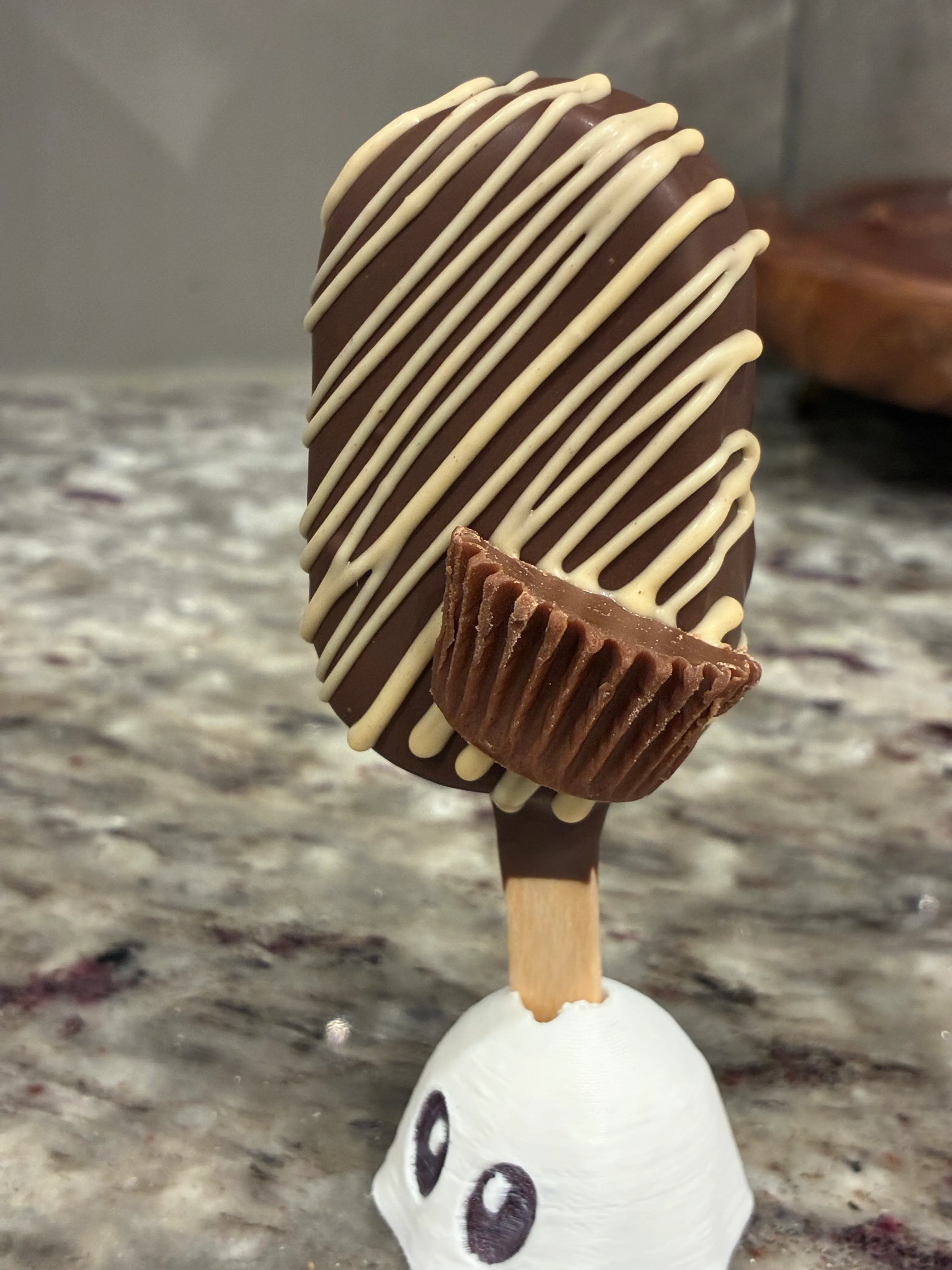 Reese’s cakesicle in a chocolate shell with white drizzle, topped with a peanut butter cup