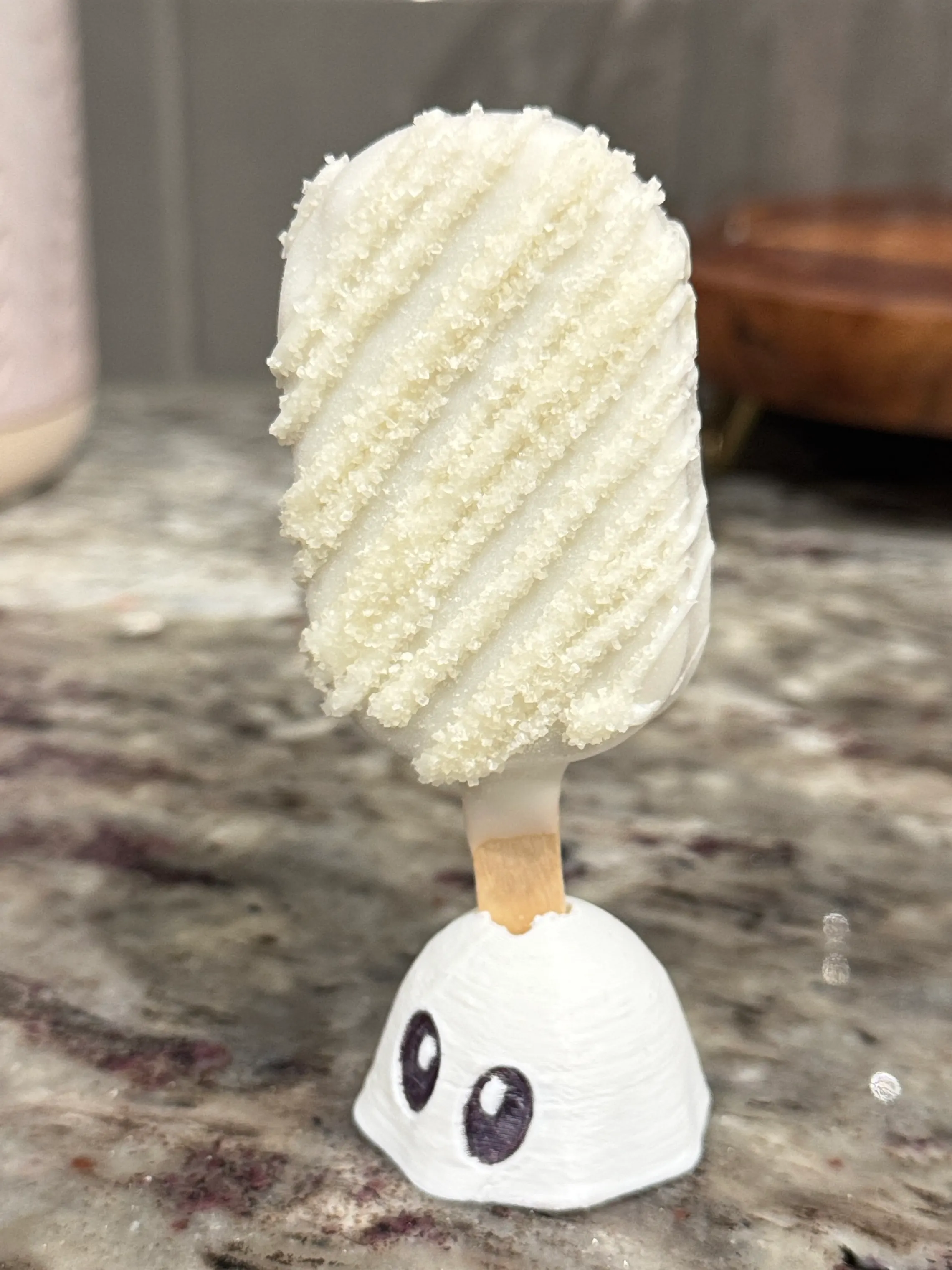 Iced sugar cookie cakesicle in a white shell with white drizzle and coarse sparkling sugar