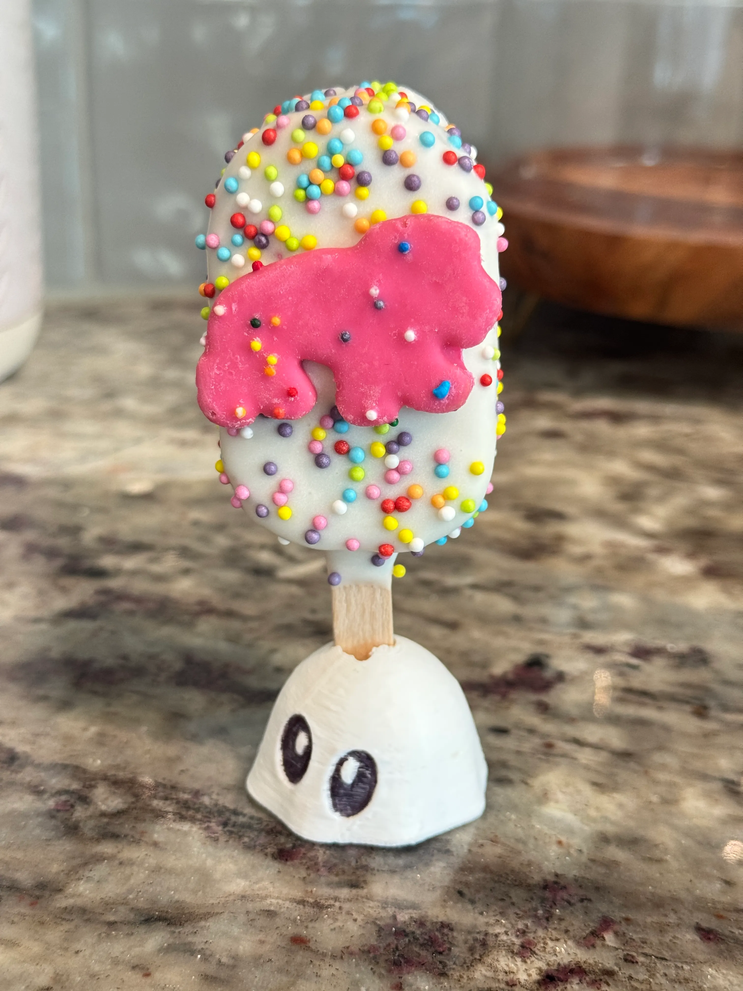 Circus cookie cakesicle in a white shell covered in rainbow nonpareils, topped with a pink circus animal cookie